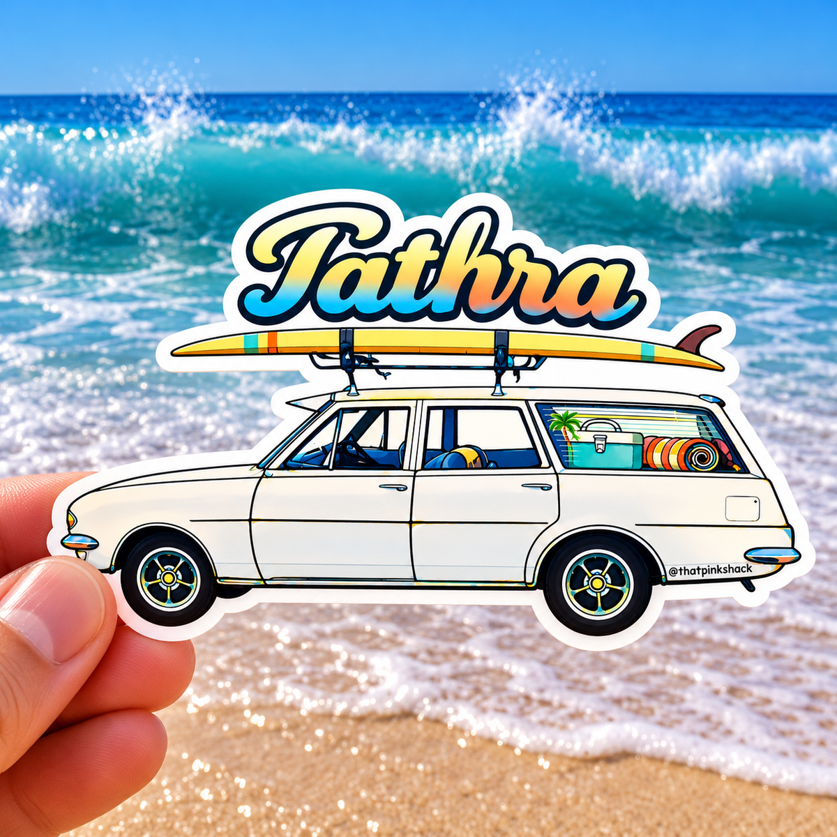 “Tathra retro surf station wagon sticker featuring a surfboard and colourful beach gear, photographed against a bright ocean and sandy beach background.”