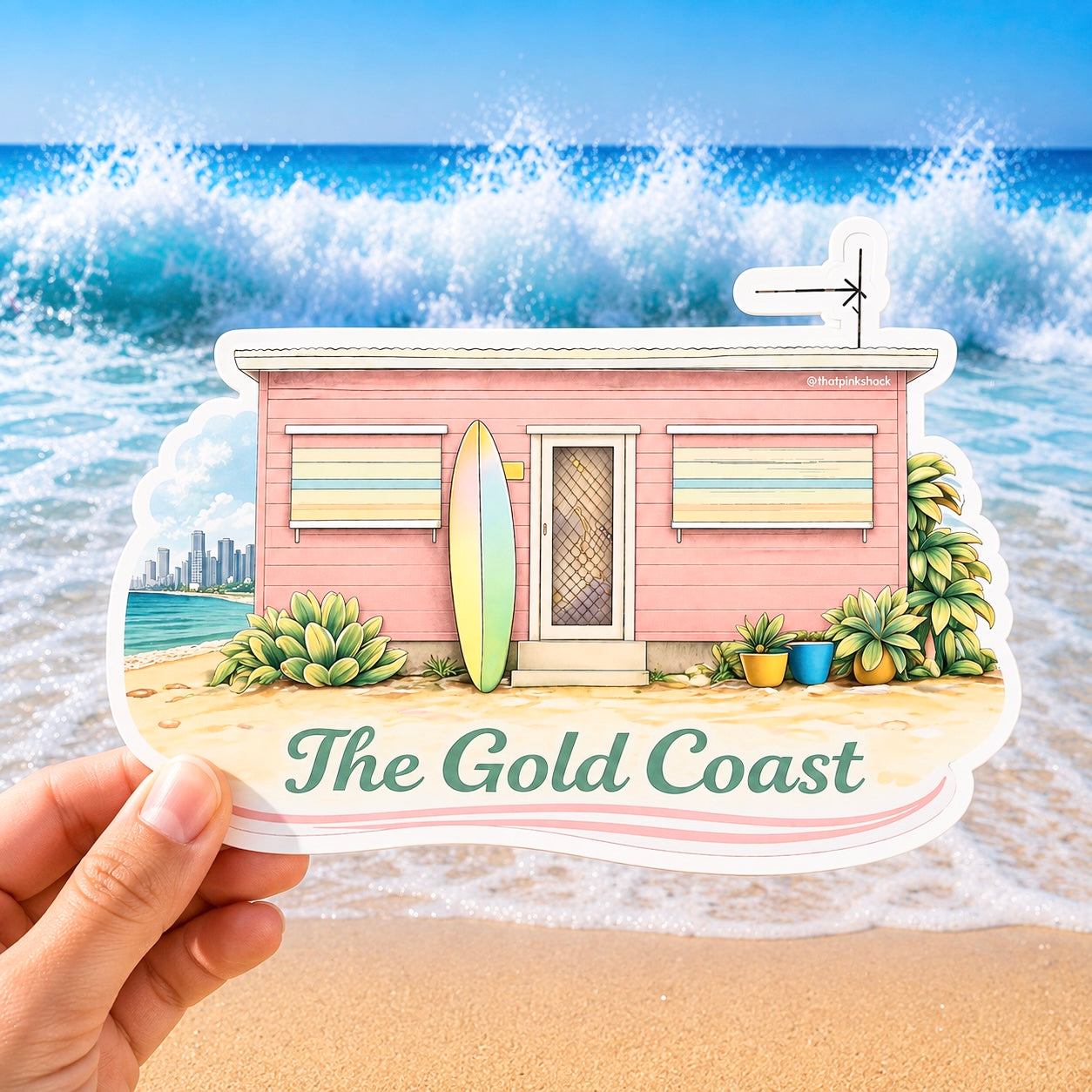 A hand holds a die-cut sticker featuring an illustrated pink beach house, surfboard, plants, and the text 'The Gold Coast' against a real beach scene with ocean waves in the background.