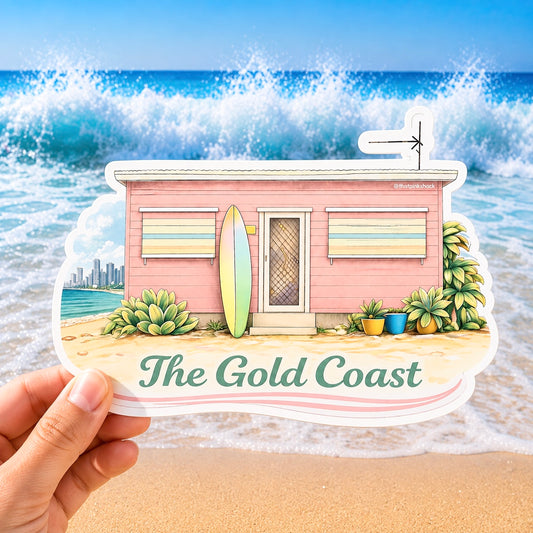 A hand holds a die-cut sticker featuring an illustrated pink beach house, surfboard, plants, and the text 'The Gold Coast' against a real beach scene with ocean waves in the background.
