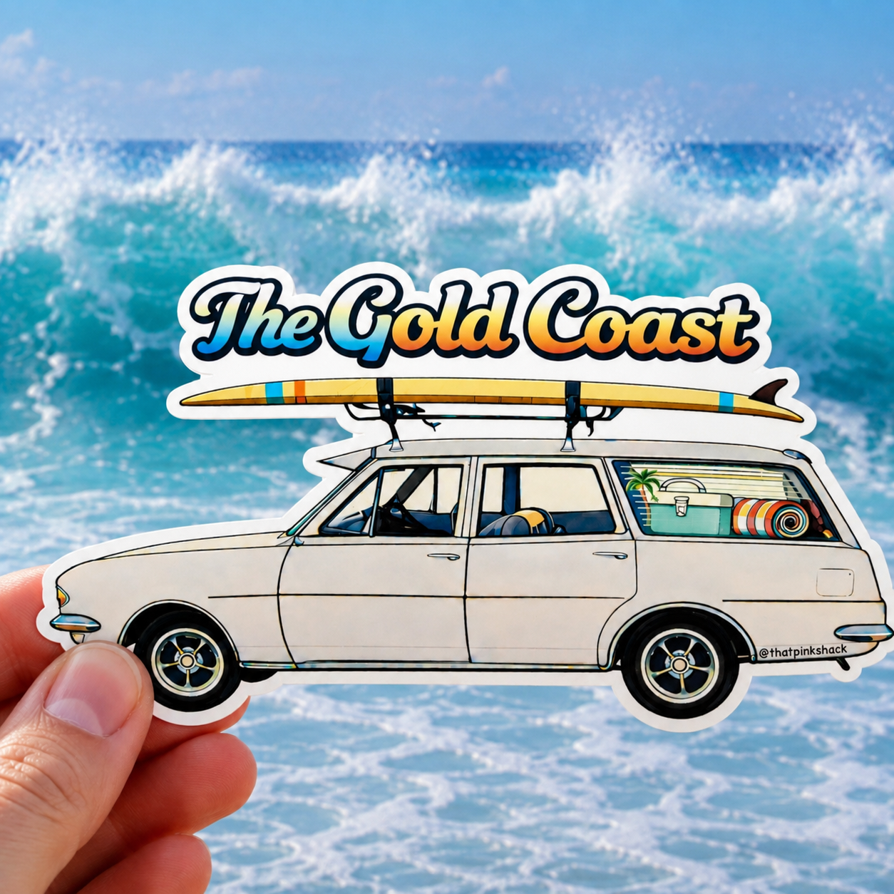 A hand holds a die-cut sticker featuring an illustrated white holden station wagon with a surfboard on its roof, the words "The Gold Coast" above, and a beach scene with ocean waves in the background.