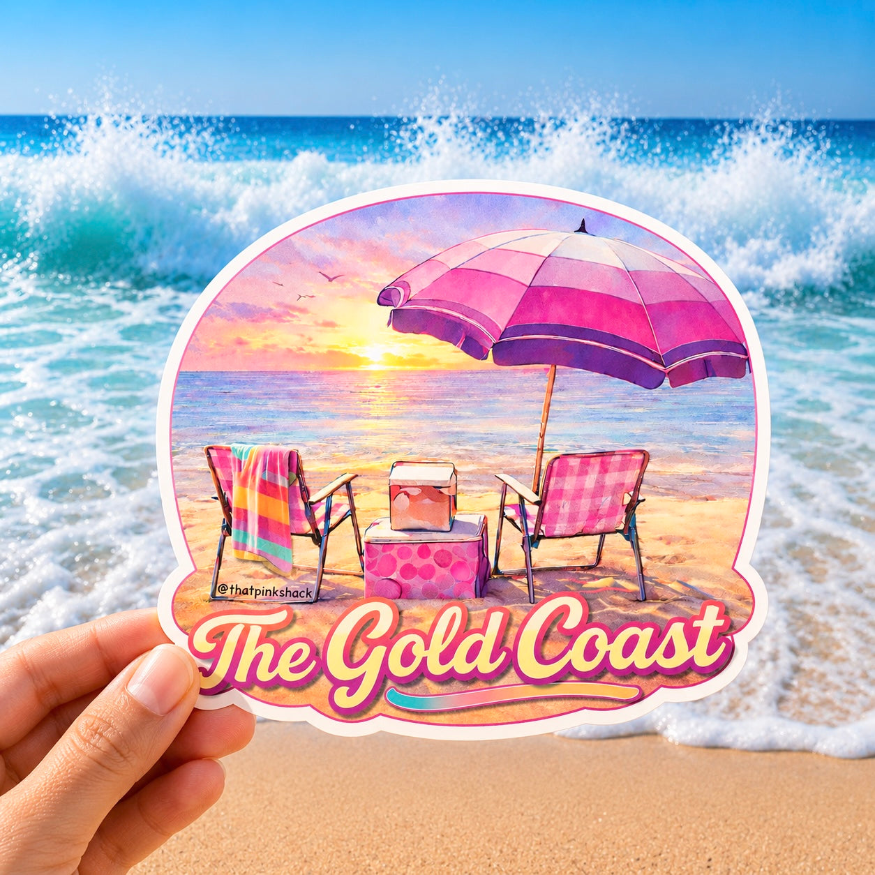 A hand holds a die-cut sticker featuring a beach scene with two lounge chairs, a pink striped umbrella, a cooler, and the text 'The Gold Coast' over a sunset. The background shows ocean waves and sandy beach.