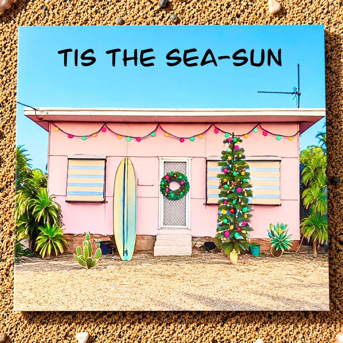 A square greeting card featuring a pastel pink beach house decorated with a green wreath, colorful string lights, a blue surfboard, and a Christmas tree, with the text 'TIS THE SEA-SUN' at the top, shown against a textured sand background.