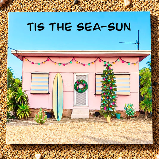A square greeting card featuring a pastel pink beach house decorated with a green wreath, colorful string lights, a blue surfboard, and a Christmas tree, with the text 'TIS THE SEA-SUN' at the top, shown against a textured sand background.