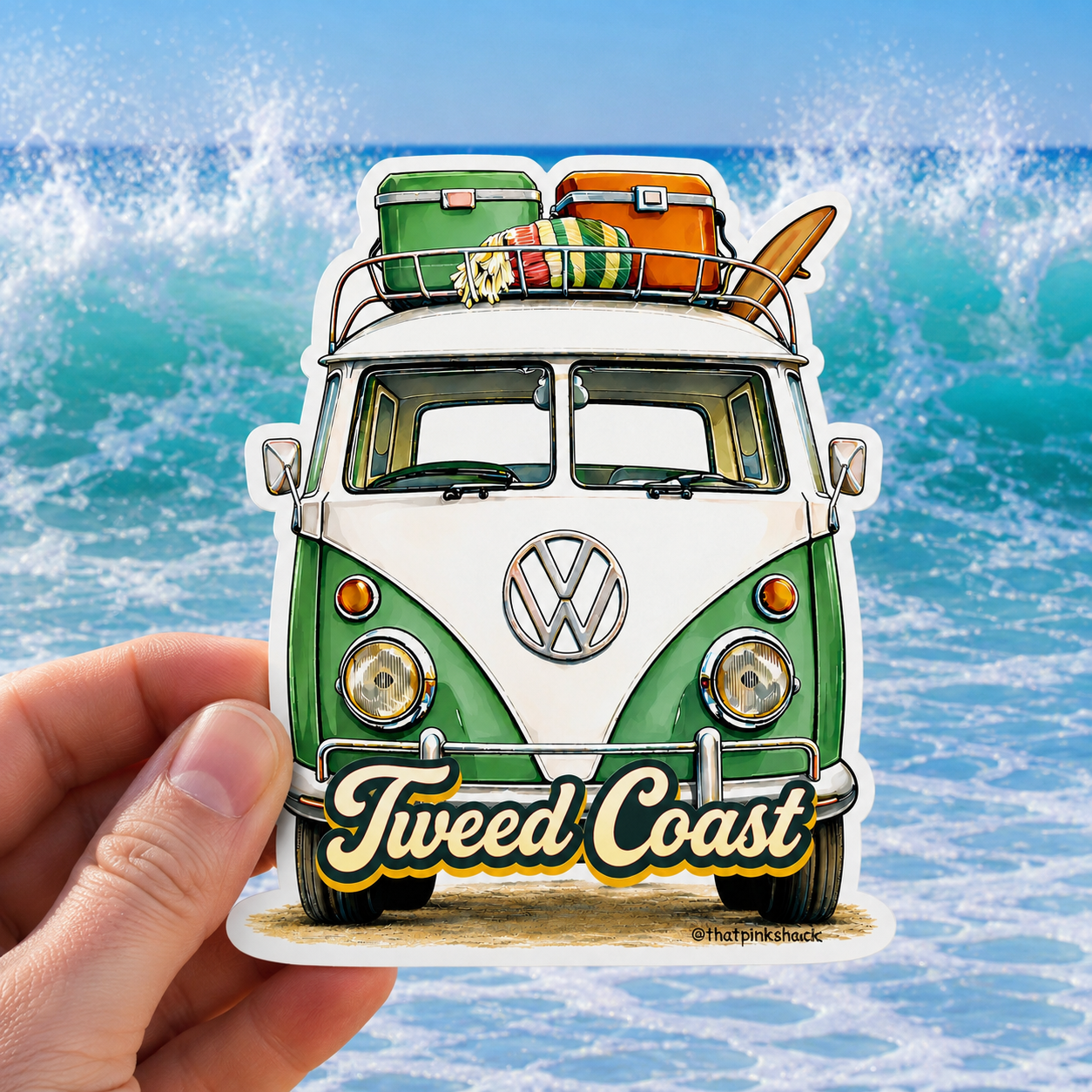 A hand holds a die-cut sticker featuring a retro green and white Volkswagen camper van with surf gear on top and the words 'Tweed Coast' on the front, set against a beach wave background.