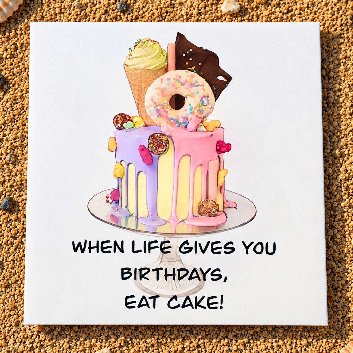 A square greeting card featuring a colorful illustration of a donut cake with the text 'WHEN LIFE GIVES YOU BIRTHDAYS, EAT CAKE!' in black capital letters, displayed on a bed of sand