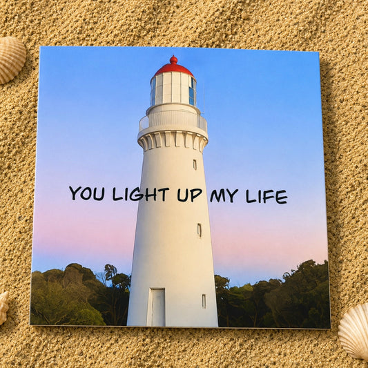 A square greeting card  featuring a white lighthouse under a blue and pink sky with the text 'YOU LIGHT UP MY LIFE', displayed on a sandy surface with seashells.