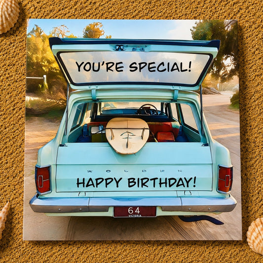 A square birthday card featuring a retro-style rear view of a light blue station wagon with its trunk open, showing a surfboard and the text 'YOU'RE SPECIAL! HAPPY BIRTHDAY!'. The card is centered on a textured sand background with small shell illustrations at the corners.