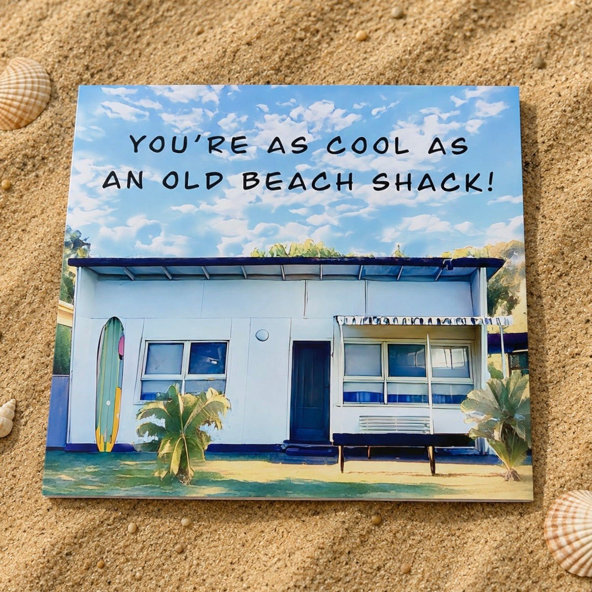 A square greeting card featuring a retro beach shack illustration and the text 'YOU'RE AS COOL AS AN OLD BEACH SHACK!' lies flat on a sandy surface with seashells nearby.