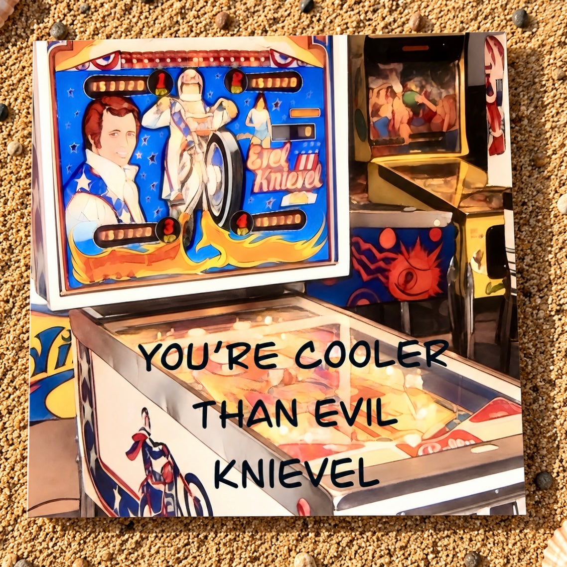 A square greeting card featuring a stylized pinball machine scene with the text 'YOU'RE COOLER THAN EVIL Knievel' overlaid in black. The artwork shows a blue playboard with racing graphics and a figure in a white suit, surrounded by sandy ground and another machine in the background.