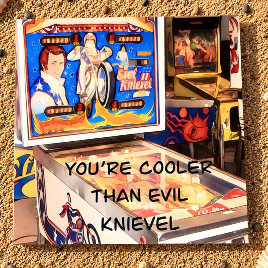 A square greeting card featuring a stylized pinball machine scene with the text 'YOU'RE COOLER THAN EVIL Knievel' overlaid in black. The artwork shows a blue playboard with racing graphics and a figure in a white suit, surrounded by sandy ground and another machine in the background.