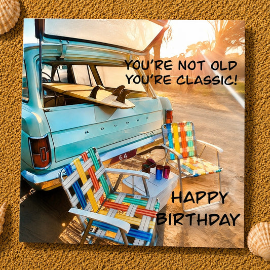 A square birthday greeting card featuring a retro scene of a turquoise vintage car trunk with surfboards, colorful striped beach chairs, and warm sunset lighting. Black text reads 'YOU'RE NOT OLD YOU'RE CLASSIC!' and 'HAPPY BIRTHDAY.