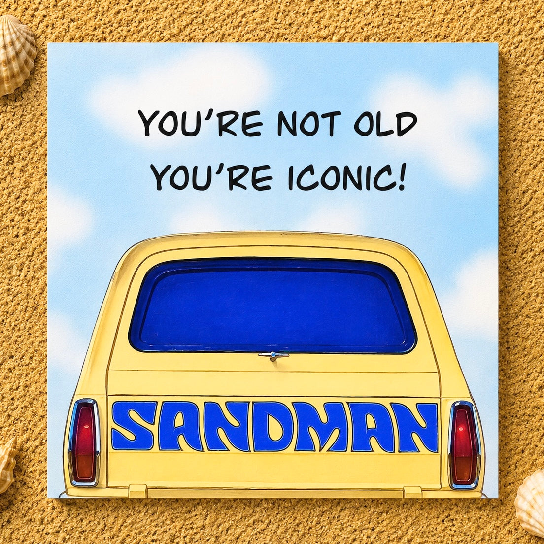 A square printed greeting card or featuring a yellow car with blue lettering reading 'SANDMAN' and the phrase 'YOU'RE NOT OLD YOU'RE ICONIC!' on a blue sky background, placed on a sandy surface with small seashells at the corners.