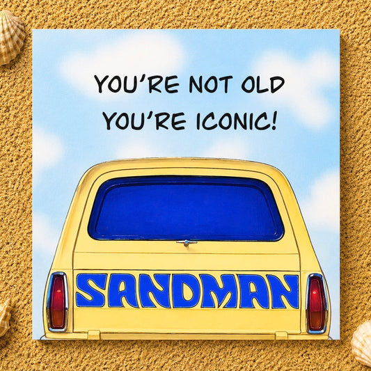 A square printed greeting card or featuring a yellow car with blue lettering reading 'SANDMAN' and the phrase 'YOU'RE NOT OLD YOU'RE ICONIC!' on a blue sky background, placed on a sandy surface with small seashells at the corners.