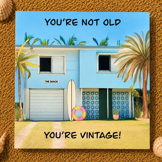 A square greeting card featuring a retro-style illustration of a light blue beach house with palm trees, a surfboard, a life preserver, and the text 'YOU'RE NOT OLD' at the top and 'YOU'RE VINTAGE!' at the bottom. The card is displayed on a textured sand background with small seashell decorations at the corners.